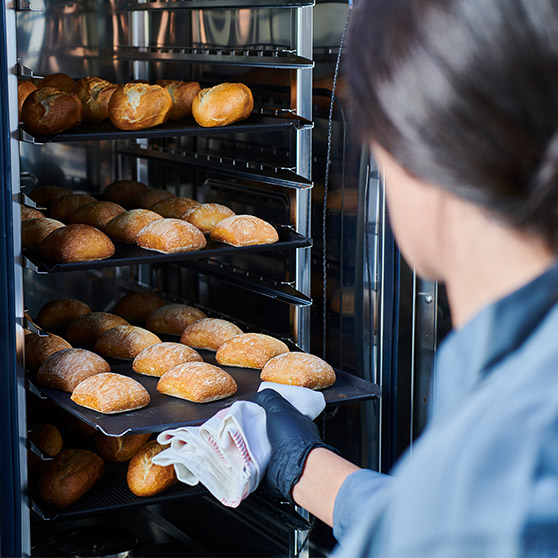 Una persona saca panecillos recién horneados del CHEF'S COMBI de Hobart usando guantes y un paño – varias bandejas con panecillos dorados en un horno profesional.