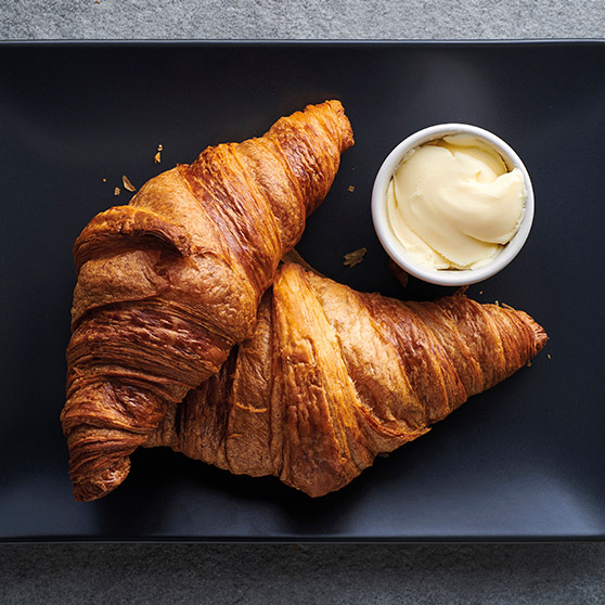Dos croissants dorados recién horneados sobre un plato negro con un cuenco de mantequilla – preparados con el CHEF'S COMBI de HOBART para una textura y dorado perfectos.