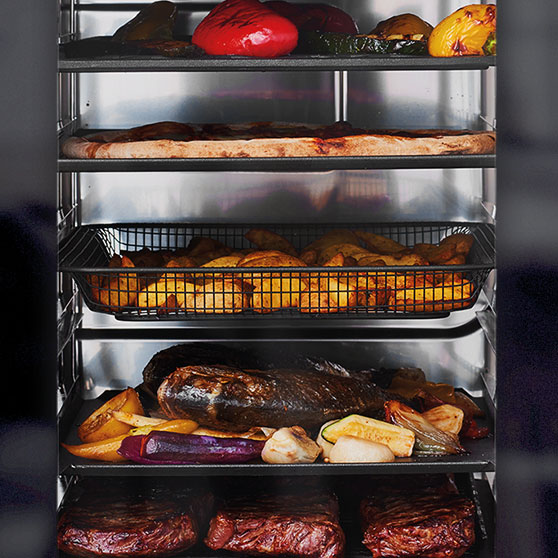 Vista interior de un horno CHEF'S KOMBI de HOBART con varios niveles – con verduras asadas, pizza, papas al horno, carne asada con vegetales y filetes, todo cocinado de forma simultánea y uniforme.