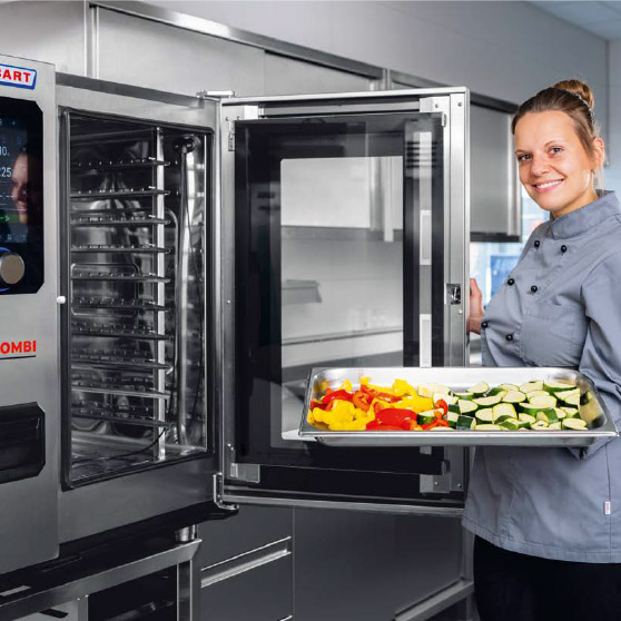 Una persona con chaqueta de chef sostiene una bandeja con verduras cortadas como pimientos y calabacín frente a un horno CHEF'S COMBI abierto con varios niveles y panel de control digital.