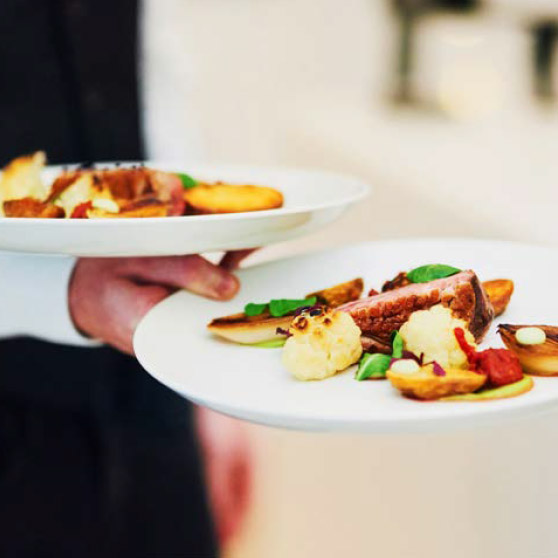 Una persona sostiene dos platos elegantemente presentados con carne, verduras asadas y guarniciones refinadas – preparados con el CHEF'S COMBI de HOBART para los más altos estándares culinarios profesionales.