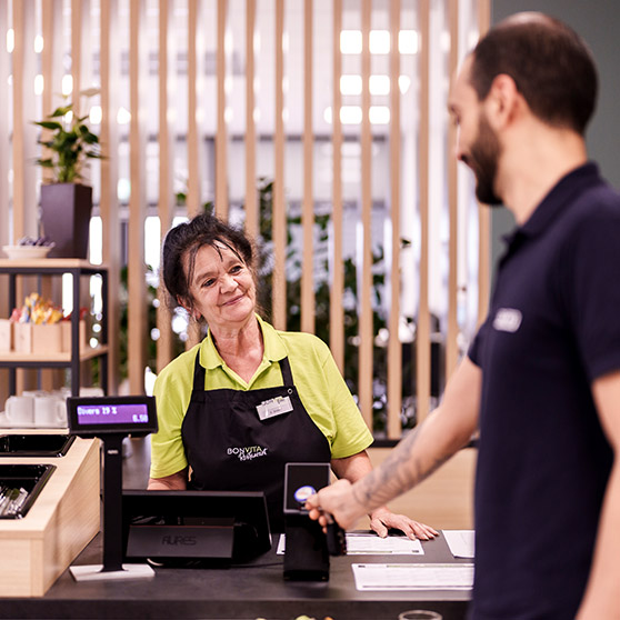 Proceso de pago en un comedor empresarial moderno: Un cliente realiza un pago sin contacto con tarjeta en un terminal electrónico. El importe aparece en una pantalla digital. Listones de madera y plantas en el fondo aportan un ambiente acogedor.