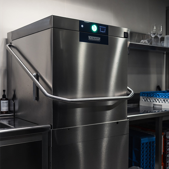 Vista lateral del lavavajillas de capota HOBART AUP en una zona de lavado profesional, acompañado de superficies y estanterías de acero inoxidable, mostrando su construcción robusta, funcionamiento silencioso y eficiencia durante picos de trabajo dentro del sistema Creatus.