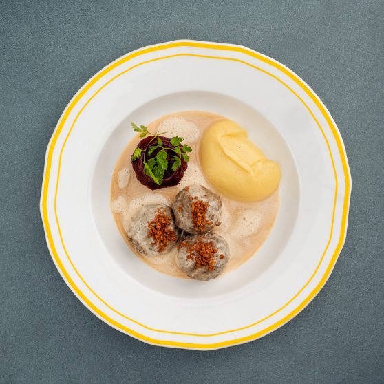 Plato blanco con borde amarillo sobre mesa gris, con albóndigas Königsberger en salsa cremosa clara y guarnición, presentación clásica.