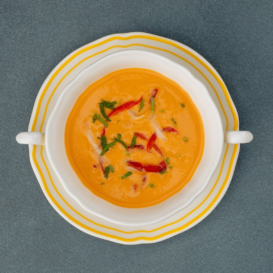 Taza de sopa blanca con borde amarillo sobre un plato, llena de sopa cremosa de color naranja. La taza tiene dos asas y está sobre una mesa gris.