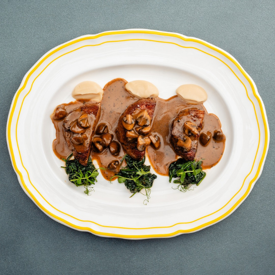 Plato blanco con borde amarillo con tres piezas de carne en salsa cremosa de champiñones, decoradas con champiñones, puré claro y hierbas verdes, presentado de forma elegante sobre mesa gris.