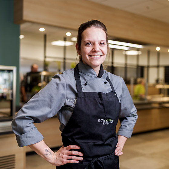 Empleada con chaqueta de cocina y delantal en una cocina empresarial moderna. Al fondo se ven mostradores y muebles de acero inoxidable.