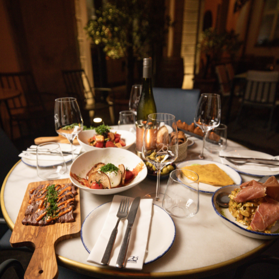 Mesa de restaurante al aire libre cuidadosamente preparada con varios platos, aperitivos, pan, una botella de vino y varias copas.