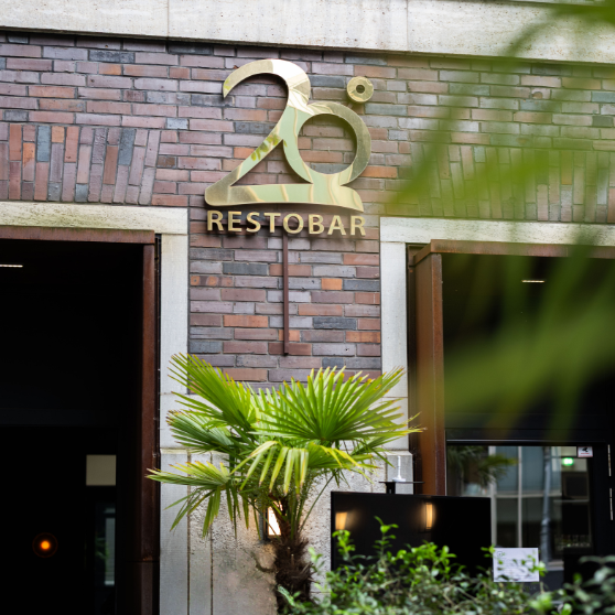 Vista exterior de un edificio de ladrillo con el rótulo dorado 20° RESTOBAR sobre la entrada, enmarcado por plantas verdes.