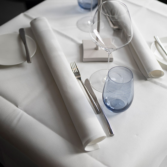 Mesa de restaurante elegantemente puesta con cubiertos alineados, copas de vino, servilletas enrolladas y un vaso azul que aportan un ambiente sofisticado.