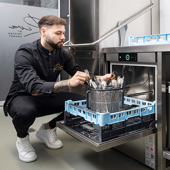 Empleado colocando cubiertos brillantes en la cesta rotatoria BESTECK PREMIUM de un lavavajillas HOBART bajo mostrador para una limpieza impecable.