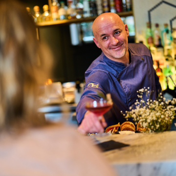 Barman sirviendo una bebida a un cliente sobre la barra, con botellas y estanterías del bar al fondo.