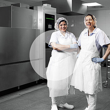 Dos empleados con ropa higiénica en la zona de lavado del hospital de Heinsberg frente a una lavavajillas industrial con carros de acero inoxidable y tecnología moderna para limpieza eficiente